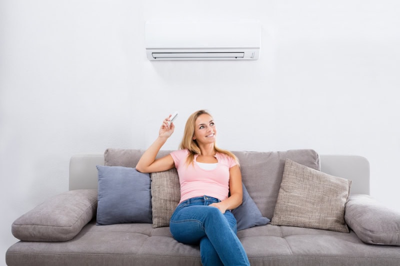 Happy woman sitting on couch operating her ductless air conditioner with a remote.