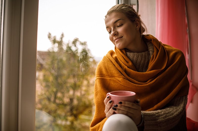 Young girl enjoying a cup of hot chocolate while sitting by window in her home.